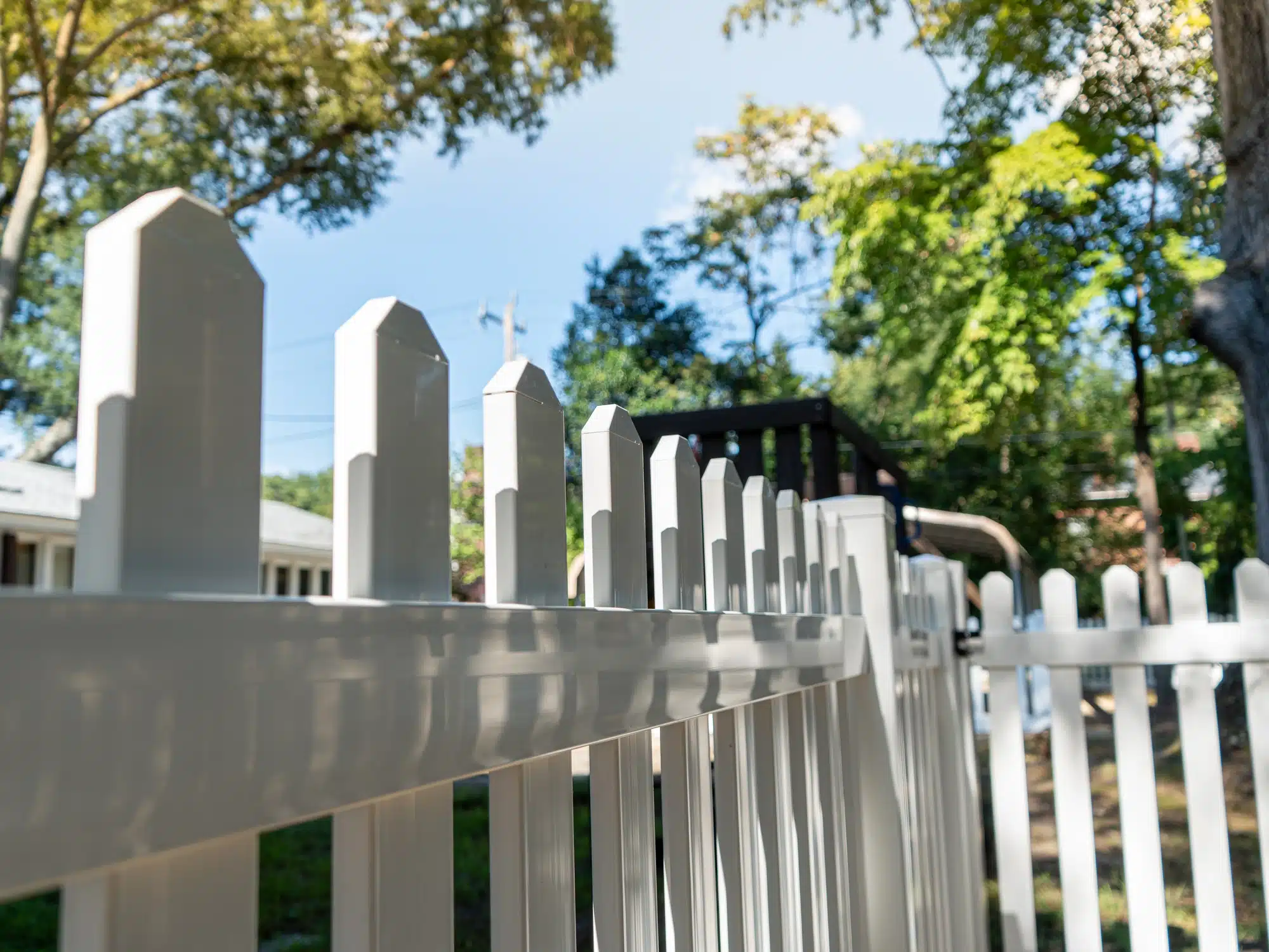 White vinyl picket fence with clipped dog-ear tops along a partly wooded yard